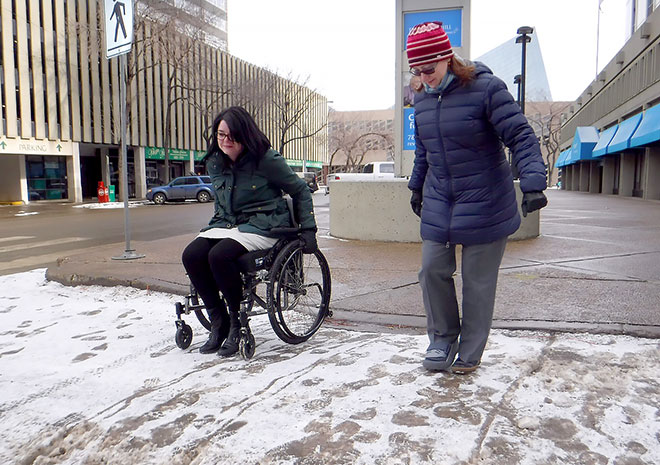 Two people crossing a snowy street, one on foot and the other in a wheelchair.