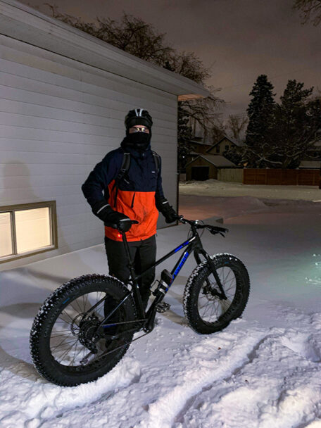 A person dressed in winter gear stand next to his bike with fat tires.