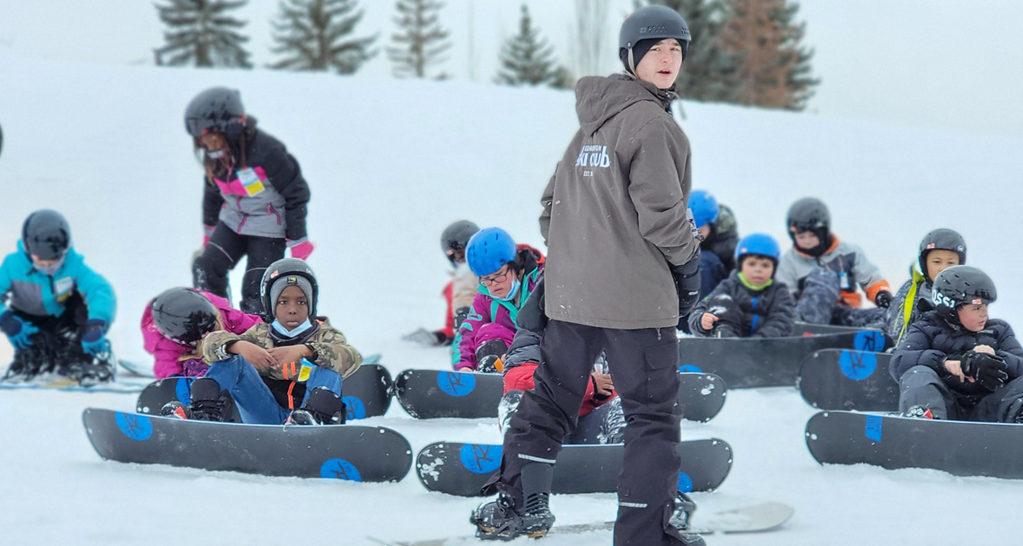 Group of kids on a snowy hill getting ready to snowboard.