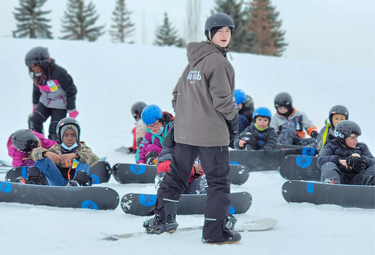 Group of kids on a snowy hill getting ready to snowboard.