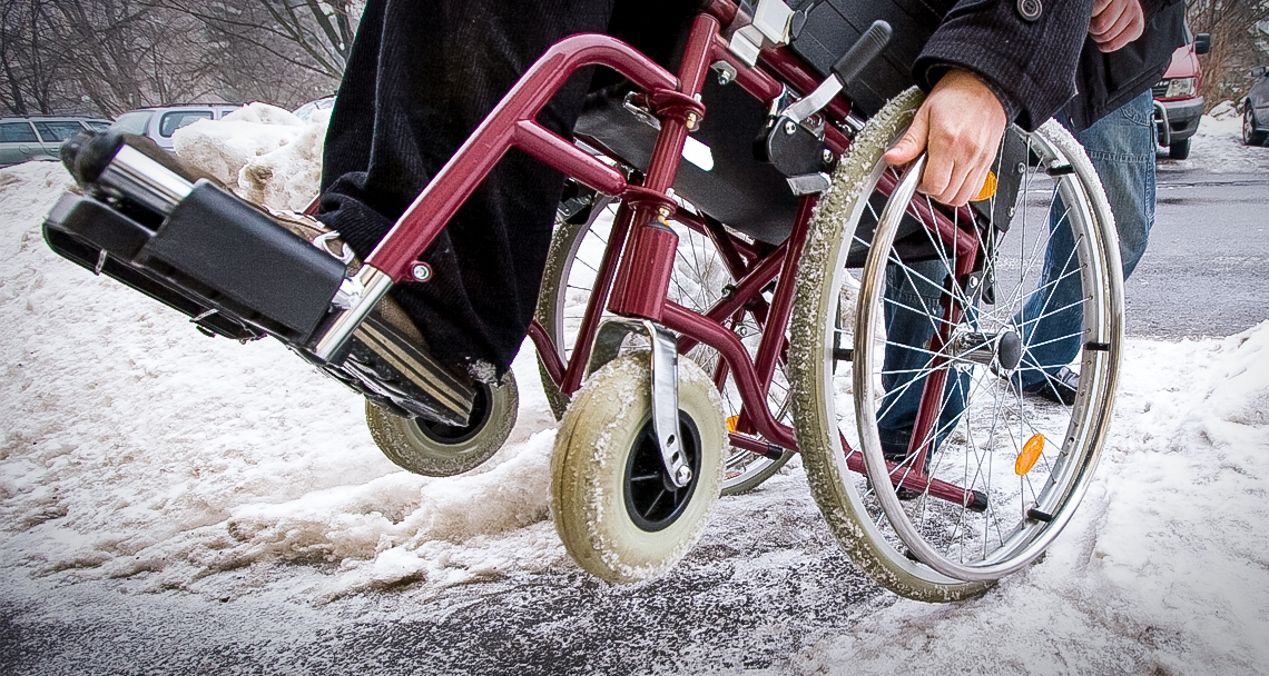 Wheelchair rider having difficulty navigating a snowy sidewalk.