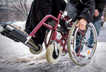 Wheelchair rider having difficulty navigating a snowy sidewalk.