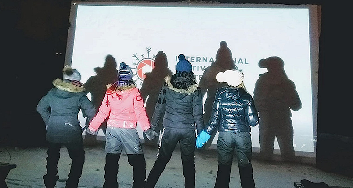 Children in snowsuits stand in front of an outdoor movie screen