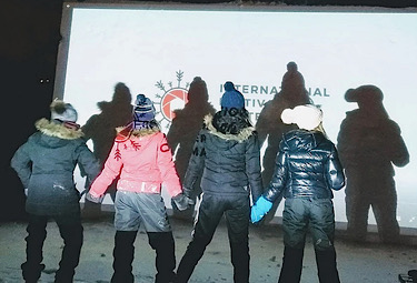 Children in snowsuits stand in front of an outdoor movie screen
